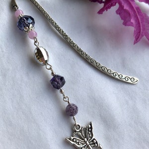 May include: A silver bookmark with a decorative design and a dangling charm featuring a silver butterfly with purple and pink beads.