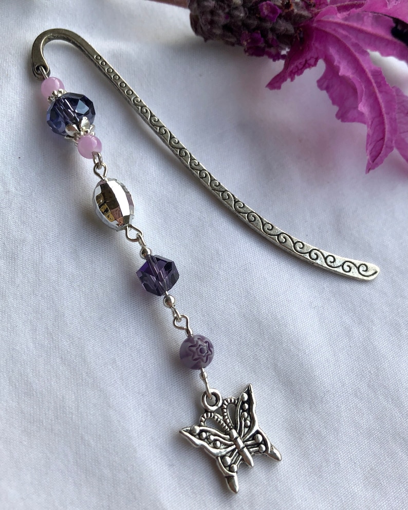 May include: A silver bookmark with a decorative design and a dangling charm featuring a silver butterfly with purple and pink beads.