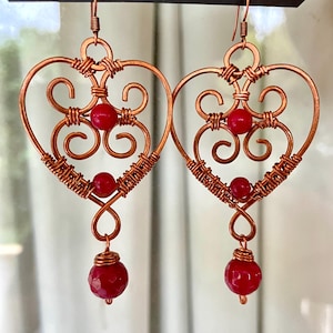May include: Copper wire heart-shaped earrings with red beads. The earrings feature intricate wire wrapping and decorative swirls. A faceted red bead dangles from the bottom of each earring, adding a pop of colour. The earrings are designed for pierced ears.