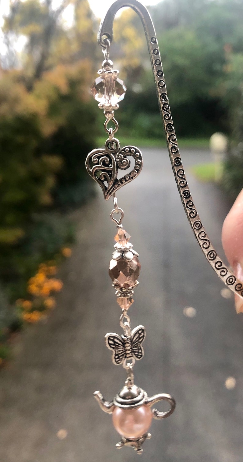 May include: A silver bookmark with a delicate heart charm, a pink crystal bead, a butterfly charm, and a teapot charm with a pink pearl. The bookmark is decorated with a repeating pattern of circles.