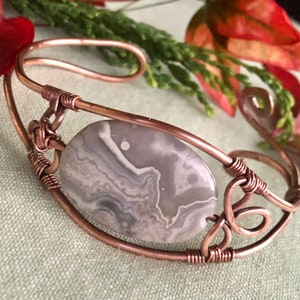 K&ouml;nnte beinhalten: Ein Armband aus Kupferdraht mit einem grauen und wei&szlig;en Achat-Stein. Der Stein ist in der Mitte des Armbands platziert und von Kupferdraht umgeben.