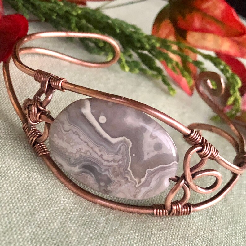 K&ouml;nnte beinhalten: Ein Armband aus Kupferdraht mit einem grauen und wei&szlig;en Achat-Stein. Der Stein ist in der Mitte des Armbands platziert und von Kupferdraht umgeben.