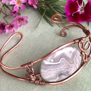 K&ouml;nnte beinhalten: Ein Armband aus Kupferdraht mit einem grauen und wei&szlig;en Achat-Stein. Das Armband hat ein zartes, filigranes Design mit einem Spiralmuster.