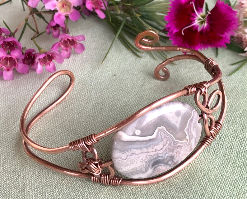 K&ouml;nnte beinhalten: Ein Armband aus Kupferdraht mit einem grauen und wei&szlig;en Achat-Stein. Das Armband hat ein zartes, filigranes Design mit einem Spiralmuster.