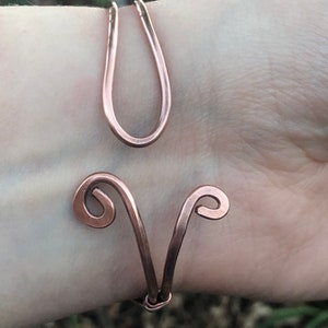 K&ouml;nnte beinhalten: Ein Armband aus Kupferdraht mit einem einzigartigen Design, das zwei geschwungene Schleifen und eine einzelne, gr&ouml;&szlig;ere Schleife aufweist. Das Armband wird am Handgelenk einer Person gezeigt.