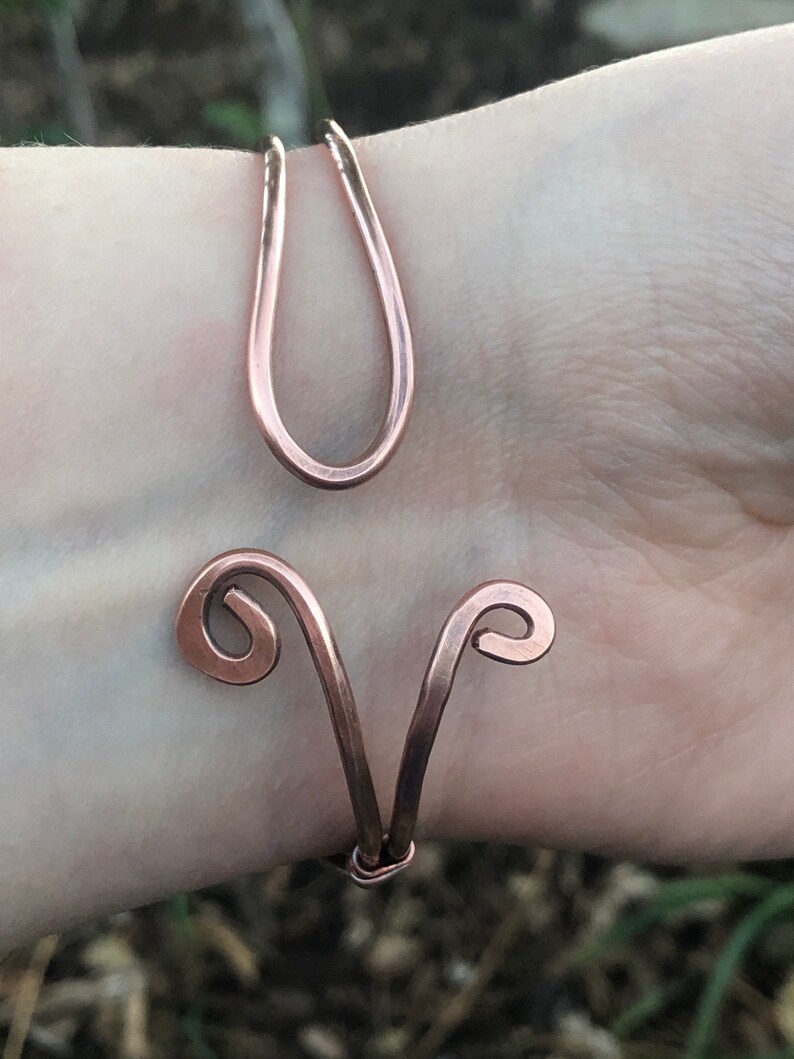K&ouml;nnte beinhalten: Ein Armband aus Kupferdraht mit einem einzigartigen Design, das zwei geschwungene Schleifen und eine einzelne, gr&ouml;&szlig;ere Schleife aufweist. Das Armband wird am Handgelenk einer Person gezeigt.