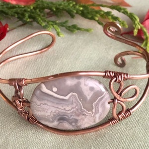 K&ouml;nnte beinhalten: Ein Armband aus Kupferdraht mit einem grauen und wei&szlig;en Achat-Stein. Das Armband hat ein zartes, filigranes Design mit einem Wirbel am Ende.