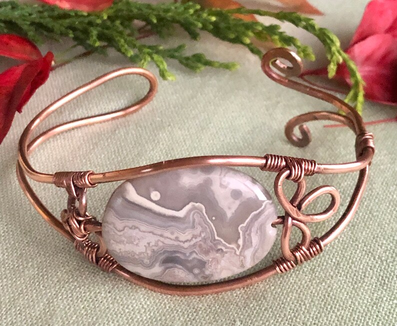 K&ouml;nnte beinhalten: Ein Armband aus Kupferdraht mit einem grauen und wei&szlig;en Achat-Stein. Das Armband hat ein zartes, filigranes Design mit einem Wirbel am Ende.