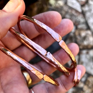 May include: Three hammered copper bangles stacked on top of each other. The bangles are a warm rose gold colour and have a slightly irregular shape.