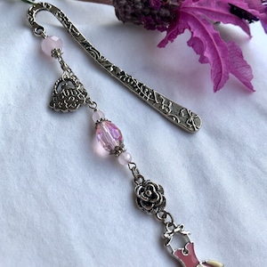 May include: A silver bookmark with a delicate floral design and a dangling charm featuring a pink dress, a rose, a crystal bead, and a heart. The bookmark is adorned with pink beads.