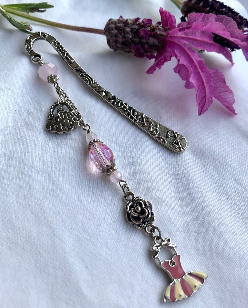 May include: A silver bookmark with a delicate floral design and a dangling charm featuring a pink dress, a rose, a crystal bead, and a heart. The bookmark is adorned with pink beads.