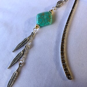 May include: Silver bookmark with a turquoise stone and three silver feathers dangling from a chain. The bookmark has a decorative design of circles along the edge.