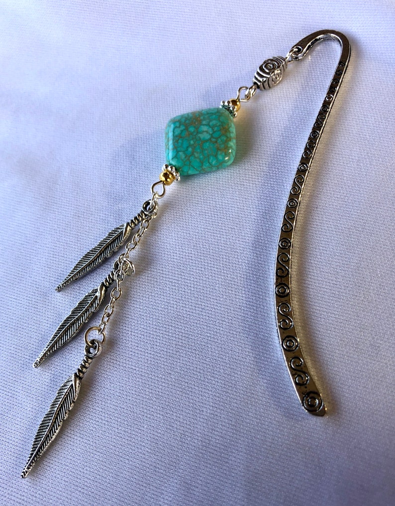 May include: Silver bookmark with a turquoise stone and three silver feathers dangling from a chain. The bookmark has a decorative design of circles along the edge.