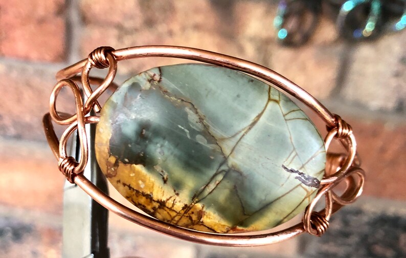 K&ouml;nnte beinhalten: Ein Armband aus Kupferdraht mit einem gr&uuml;n-braunen Achat-Stein. Der Stein ist oval geformt und hat ein nat&uuml;rliches Muster.