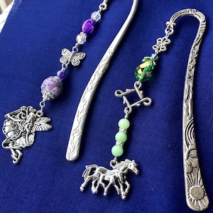 May include: Three decorative metal bookmarks with dangling charms. One features a fairy, purple beads, and a butterfly. Another has green beads and a horse charm. The third has a sun design. All are on a blue background.