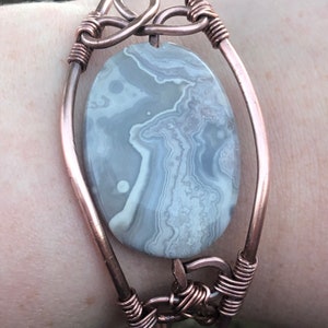 K&ouml;nnte beinhalten: Ein mit Kupferdraht umwickeltes Armband mit einem grauen und wei&szlig;en Achat-Stein. Das Armband hat ein einzigartiges Design mit einem Knoten oben und unten.