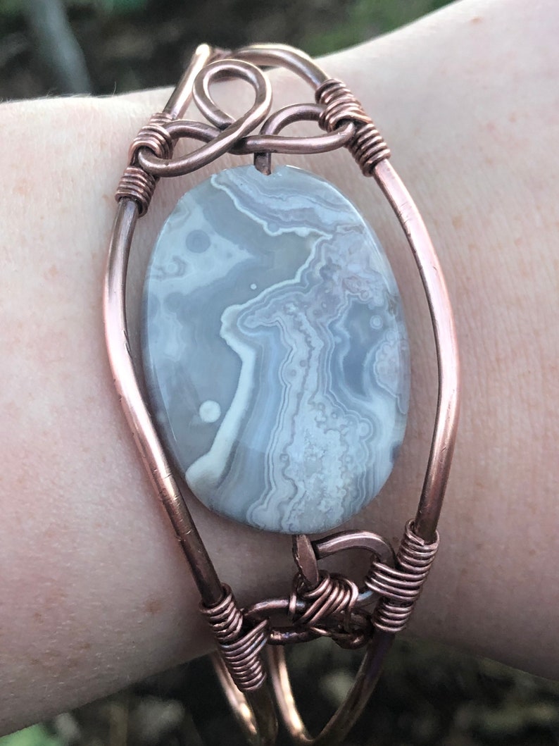 K&ouml;nnte beinhalten: Ein mit Kupferdraht umwickeltes Armband mit einem grauen und wei&szlig;en Achat-Stein. Das Armband hat ein einzigartiges Design mit einem Knoten oben und unten.