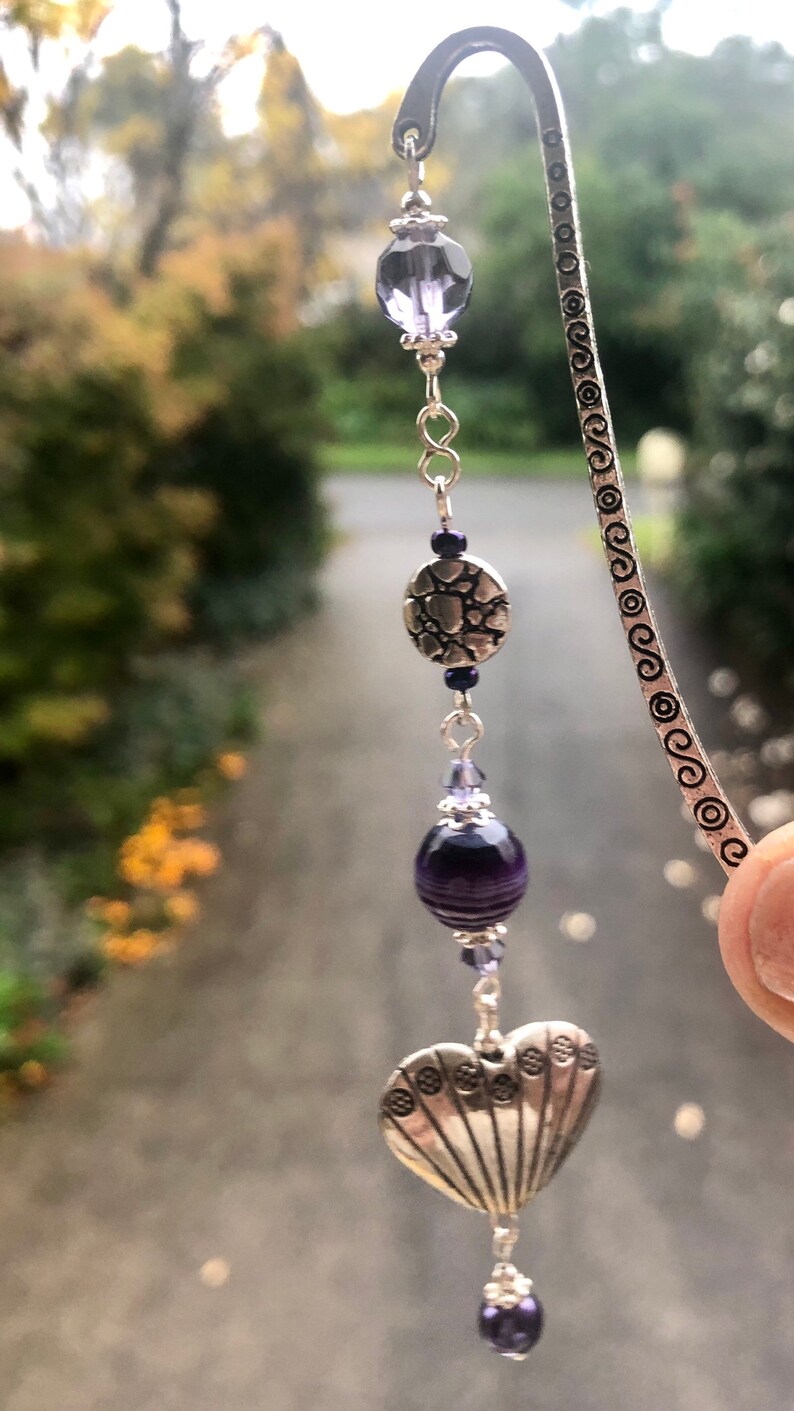 May include: A silver bookmark with a heart-shaped charm, a round charm with a textured design, and purple glass beads.