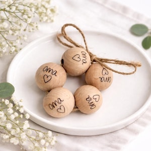 Könnte beinhalten: Fünf Holzperlen mit dem Wort "oma" und einem Herz, verbunden durch eine Schnur. Sie liegen auf einem weißen Teller, mit weißen Blumen und grünem Laub im Hintergrund. Die Perlen sind aus Holz.