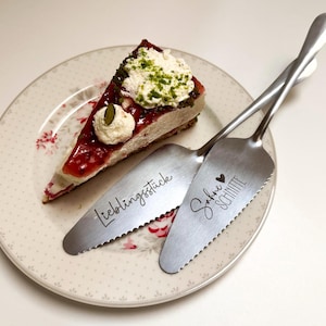 Könnte beinhalten: Ein Stück Käsekuchen mit rotem Belag und Schlagsahne auf einem Teller mit Blumenmuster, serviert mit zwei silbernen Tortenhebern. Einer trägt den Text "Lieblingsstück", der andere "Sahne Schnitte" mit einem Herzsymbol.