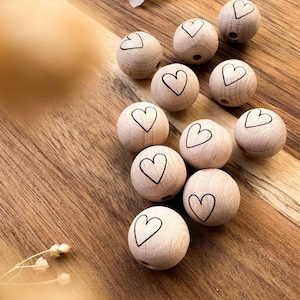 Könnte beinhalten: Ein Set aus zehn Holzperlen mit einem Herz-Design. Die Perlen sind hellbraun und haben eine glatte Oberfläche. Sie sind perfekt für die Herstellung von Schmuck oder anderen Bastelarbeiten.