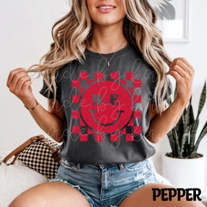 Op de afbeelding: Donkergrijs T-shirt met een rood-zwarte geblokte smiley-gezicht grafiek. De grafiek bevat de letters "NE" in het smiley-ontwerp. Het woord "PEPPER" staat in het wit onderaan de afbeelding.