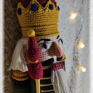 May include: A crocheted nutcracker doll with a red and yellow crown, a red and yellow staff, and a white beard. The doll is wearing a green and yellow uniform with a red bow tie. The doll is standing on a white fuzzy surface.