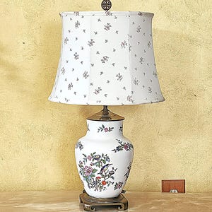 May include: A table lamp with a white ceramic base featuring colorful floral and bird patterns. The lamp has a white fabric shade with a small floral print and a dark metal finial at the top. The base rests on a square dark metal stand.