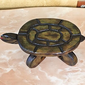 May include: A wooden turtle-shaped decorative item. The turtle has a dark green and brown shell with a black outline, a head with a red mouth, and four legs. The item is made of wood and has a rustic appearance.