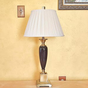 May include: A gold table lamp with a white pleated lampshade and a black ceramic base. The lamp is on a wooden table.