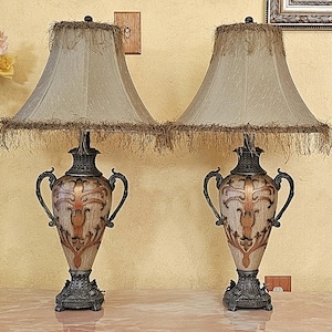 May include: A pair of table lamps featuring urn-shaped bases with dark bronze-toned metal accents and ornate copper-colored scrollwork. Each lamp has a bell-shaped fabric shade in a neutral beige tone, finished with decorative brown fringe trim.