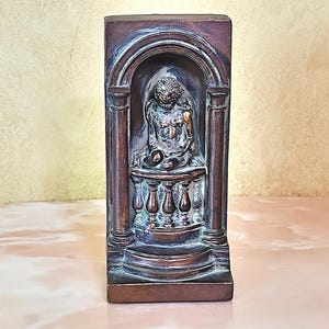 May include: A bronze-colored bookend with a sculpted figure of a person sitting on a balcony. The figure is holding a round object in their lap. The bookend has a detailed design with columns and an archway.