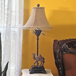 May include: A table lamp featuring a dark brown metal base shaped like a palm tree with a camel figure standing on a square pedestal. The lamp has a beige fabric lampshade with a textured weave and a decorative fringe of small clear beads.