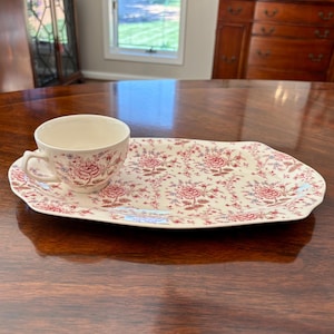 Könnte beinhalten: Ein Vintage-Teebecher- und Serviertablett-Set. Die Tasse und das Tablett sind mit einem Blumenmuster in Rot und Blau auf cremefarbenem Hintergrund verziert. Das Tablett ist rechteckig mit einem gewellten Rand.