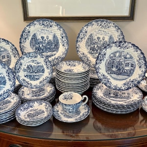 May include: A full blue and white porcelain dinner service. The set includes plates, bowls, cups, and saucers. Each piece features a detailed illustration and a scalloped edge. The tableware is arranged on a wooden surface.