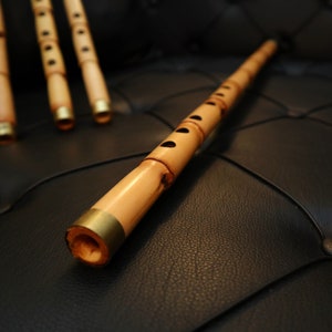 May include: Four light brown bamboo flutes with dark circular finger holes and metallic gold-coloured rings at the ends. The flutes are arranged with one positioned horizontally in the foreground and three others placed vertically in the background.