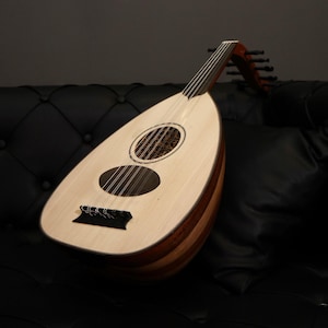 May include: A light-coloured oud, a stringed instrument, rests on a black leather surface. The pear-shaped body features a sound hole and multiple strings. The neck and tuning pegs are visible, showcasing the instrument's design.