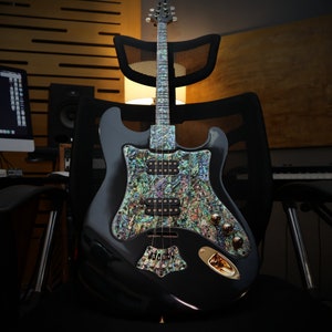May include: A black electric guitar with a pearlescent pickguard and fretboard sits on a black chair. The guitar has gold-colored knobs and a gold-colored plate. The background includes studio equipment.