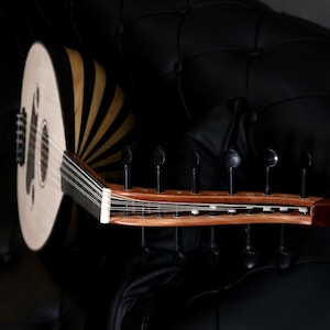 May include: An oud, a stringed instrument, rests on a black surface. The instrument features a light-coloured soundboard with dark wood accents and a wooden neck with tuning pegs. The strings are visible, and the instrument is angled.