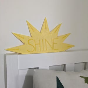 Può includere: Decorazione a forma di sole in feltro giallo con la scritta "SHINE" ricamata al centro. La decorazione è montata su una testiera bianca, con un cuscino con un disegno di albero verde sottostante.