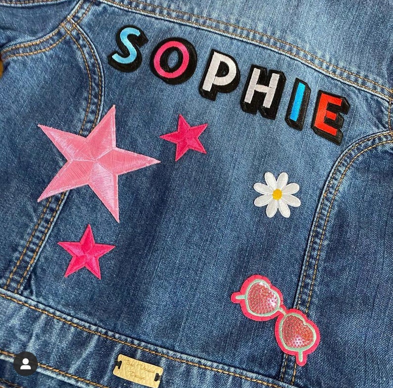 May include: A blue denim jacket with colorful patches, including the name "SOPHIE" in rainbow letters, pink stars, a white daisy, and pink sunglasses.