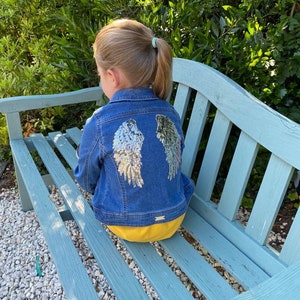 May include: A young person wearing a blue denim jacket with silver sequin angel wings on the back. The person is sitting on a light blue wooden bench.