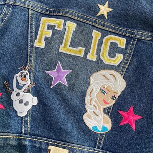 May include: A blue denim jacket with various patches, including a gold embroidered "FLIC" patch, a purple star, a pink star, a white Olaf patch, and a blue Elsa patch. The jacket also has a brown leather patch with the text "Made with Love for Your Little Darling".