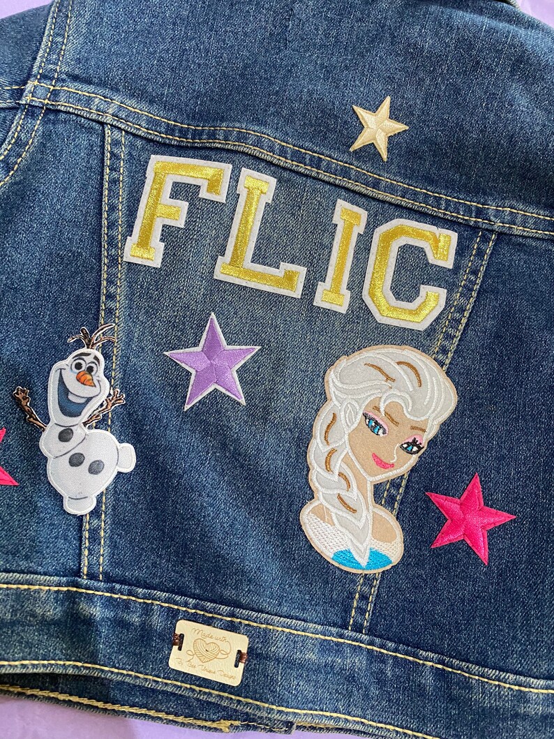 May include: A blue denim jacket with various patches, including a gold embroidered "FLIC" patch, a purple star, a pink star, a white Olaf patch, and a blue Elsa patch. The jacket also has a brown leather patch with the text "Made with Love for Your Little Darling".