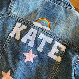 Op de afbeelding: Een blauwe denimjas met een witte viltpatch waarop "KATE" staat en een regenboog- en wolkpatch. Er zitten twee roze viltsterren op de jas.