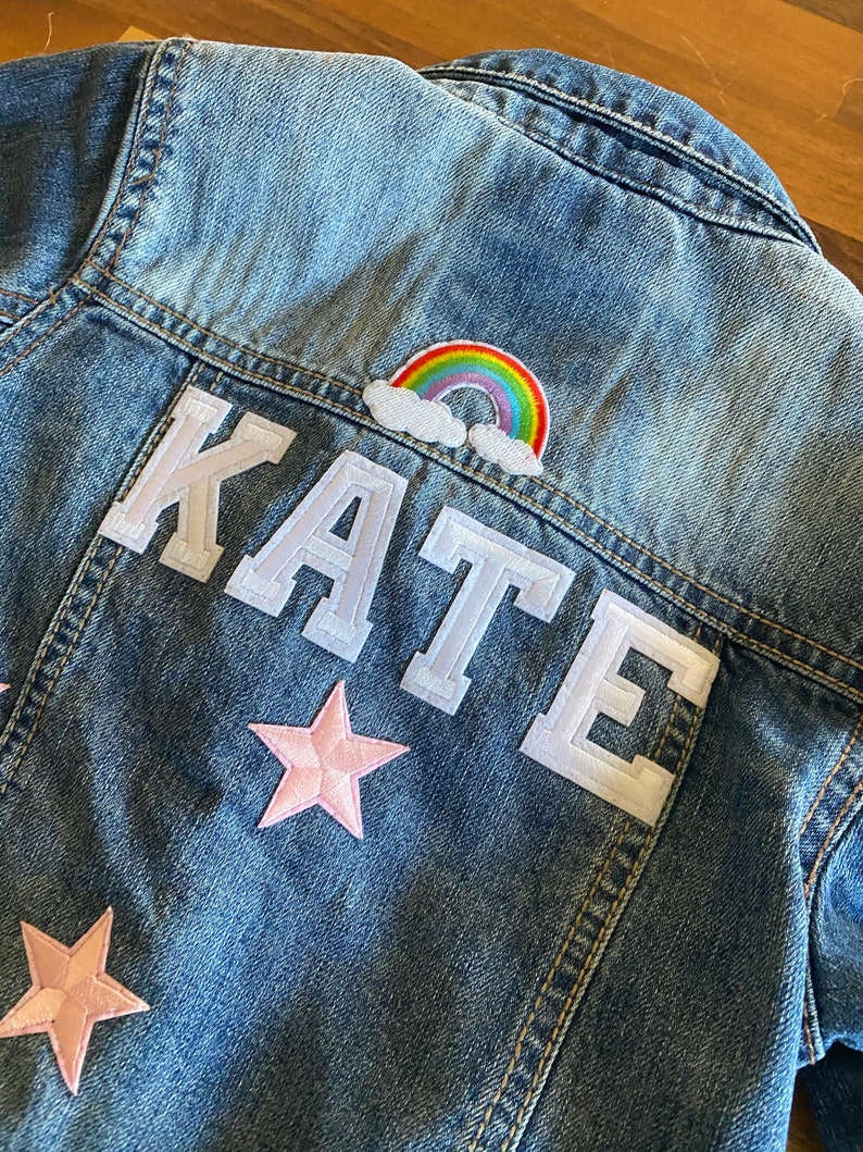 Op de afbeelding: Een blauwe denimjas met een witte viltpatch waarop "KATE" staat en een regenboog- en wolkpatch. Er zitten twee roze viltsterren op de jas.