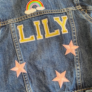 Op de afbeelding: Een blauwe denim jas met een regenboog, wolken en sterren patches. De naam "LILY" is geborduurd in gouden letters op de rug.