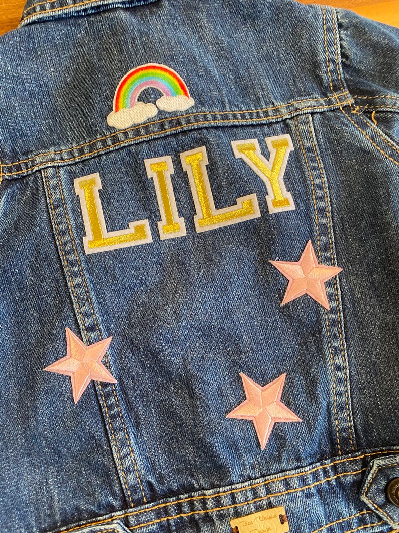 Op de afbeelding: Een blauwe denim jas met een regenboog, wolken en sterren patches. De naam "LILY" is geborduurd in gouden letters op de rug.