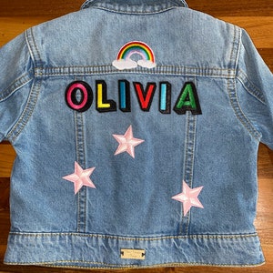 Op de afbeelding: Een lichtblauw denim jack met kleurrijke patches. De patches spellen de naam "OLIVIA" en bevatten een regenboog en roze sterren.