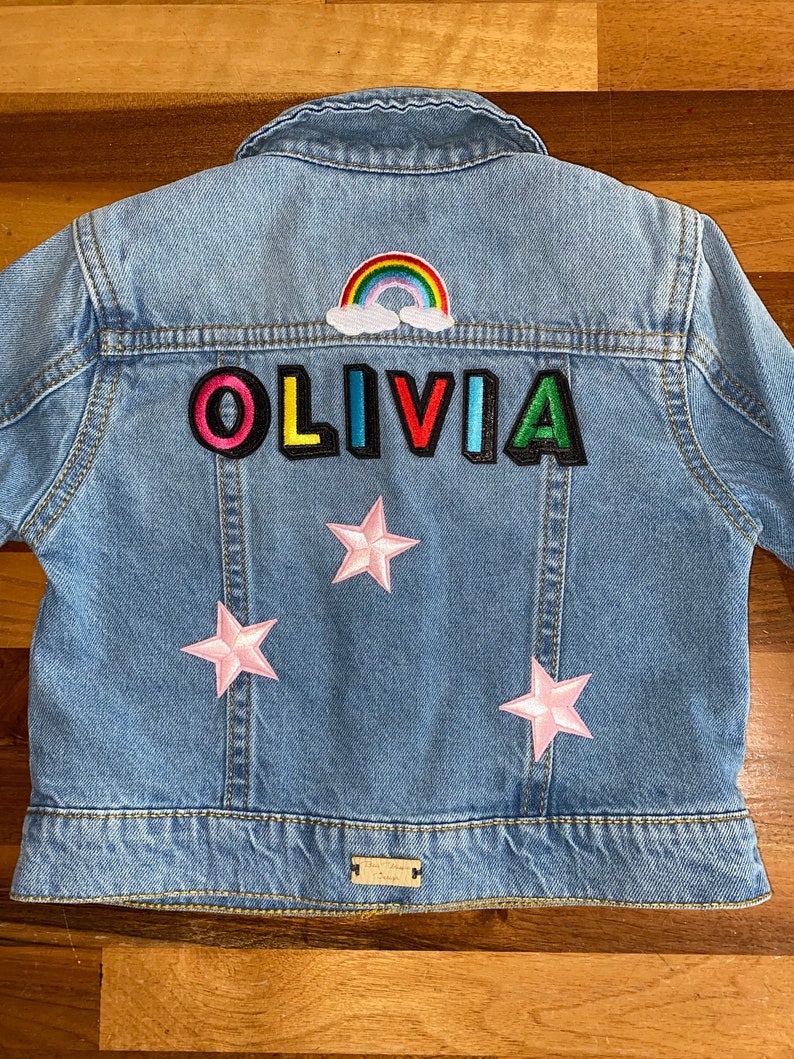 Op de afbeelding: Een lichtblauw denim jack met kleurrijke patches. De patches spellen de naam "OLIVIA" en bevatten een regenboog en roze sterren.
